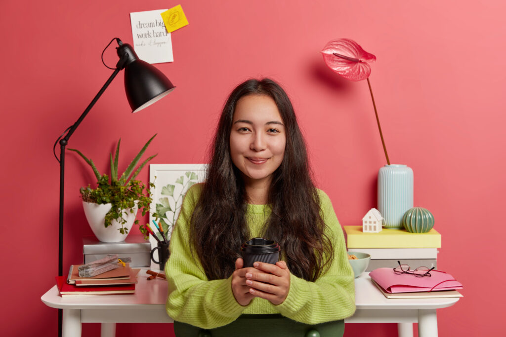 Pretty korean female scholar in green sweater, holds takeawy coffee cup, prepares research at home, poses against cozy workplace with flowers, desk lamp, textbooks and folders. Pink background