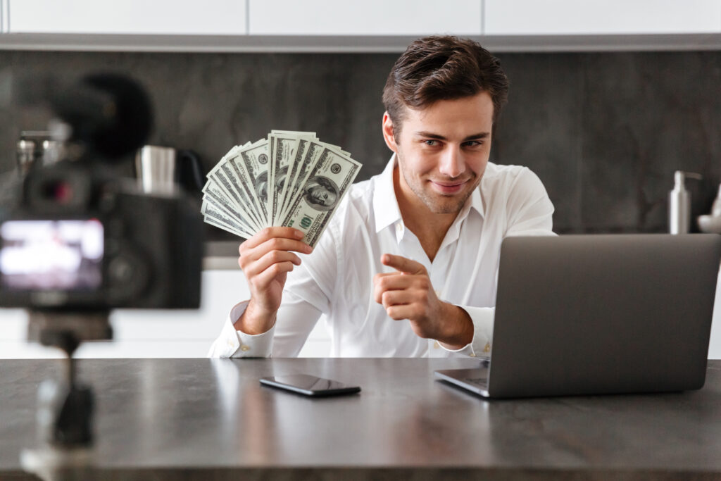 sorrindo-jovem-filmando-seu-episodio-de-blog-de-video-sobre-novos-dispositivos-de-tecnologia-enquanto-esta-sentado-na-mesa-da-cozinha-com-o-laptop-e-mostrando-um-monte-de-notas-de-dinheiro-scaled.
